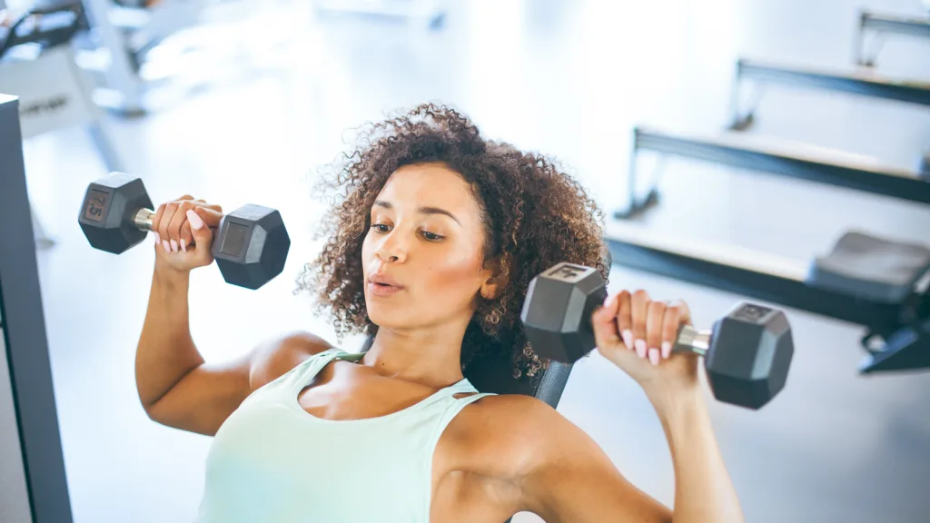 Woman lifting weights as she lays on the bench in a gym. She is wearing a tank top and has curly hair; her lips are pierced as she exhales while exercising. New pet-friendly apartment homes for rent with private fenced backyards in Arizona, Texas, Florida, and North Carolina with pool, pickleball, dog park, gym, clubhouse and gated community. *No breed or weight restrictions. Yardly